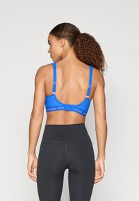 Modrá športová podprsenka s kontrastnými ružovými akcentmi; štruktúrovaná látka, nastaviteľné ramienka a široký pruh s označením "traction". Čierne vysoké legíny.