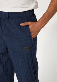 Tamsiai mėlynos, lengvos kelnės su tekstūriniu paviršiumi, elastinga juosmens juosta, šonine kišenė su užtrauktuku ir juodu "Guess" logotipo pleistru.