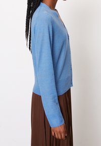 Hellblauer Cardigan aus weichem Material, mit einem runden Ausschnitt, Knopfverschluss, langen Ärmeln und gerippten Bündchen.