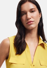 Yellow sleeveless blouse with a collar, featuring two front patch pockets and a smooth texture, paired with subtle metallic accents.
