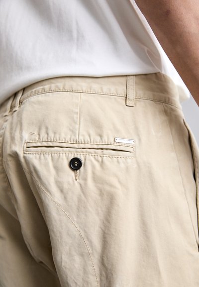 Prim-plan al unei buzunare de pantalon beige, cu un nasture negru și un mic etichet metalic de marcă, purtat cu o cămașă albă.