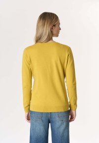 Maglione giallo senape con maniche lunghe, scollo a girocollo, bordo a costine e texture morbida, abbinato a jeans di denim azzurri chiari.