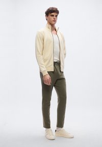 Kermanvärinen vetoketjullinen takki, harmaa hihaton paita, oliivinvihreät housut ja vaaleat lenkkarit. Sileä kangas, kapea istuvuus ja säädettävä vyötärönauha.