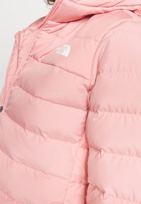 Veste rose rembourrée avec fermeture éclair et capuche. Elle possède des sections quiltées horizontales et un petit logo blanc sur la poitrine.