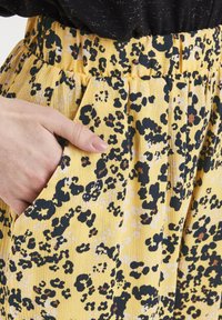 Keltainen kangas leopardikuvioin, jossa mustia, ruskeita ja valkoisia yksityiskohtia. Sisältää laskostetun vyötärökaitaleen ja sivutaskut.