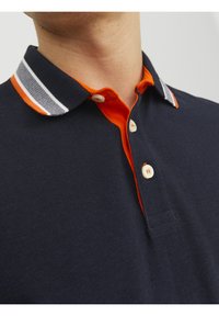 Marineblå polo med struktureret stof. Har en orange indvendig krave og stribede detaljer i hvid og grå på den ydre krave. To knapper.