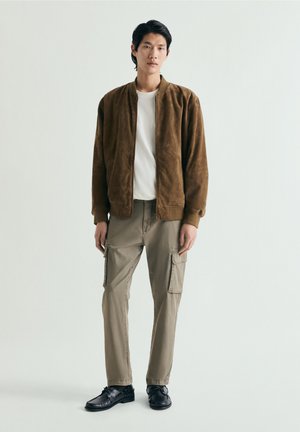 Jeune homme debout portant une veste bomber en daim marron, un t-shirt blanc, un pantalon cargo beige et des chaussures en cuir noir sur un fond uni.