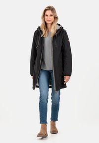 Schwarze Kapuzenparka-Jacke mit Reißverschluss, ausgestattet mit einem grauen Innenfutter und Kordelzügen. Getragen über einem grauen Oberteil und Jeans.