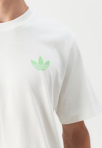 White cotton t-shirt with a small green three-stripe logo on the left chest. Short sleeves and a crew neckline. Smooth texture.