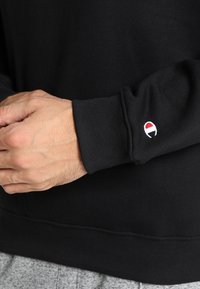 Felpa nera con una texture morbida, caratterizzata da un polsino a costine e un piccolo logo ricamato in rosso e bianco sulla manica.