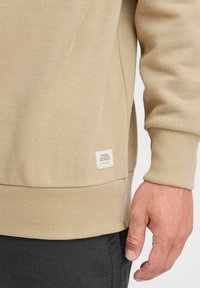 Beige sweatshirt met ribgebreide manchetten en zoom, met een klein wit merkenlabel aan de zijkant. Glad oppervlak met een losse pasvorm.