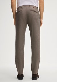 Pantaloni marroni con texture, dal taglio skinny, con tasche posteriori e una leggera lucentezza. Abbinati a scarpe scure e una maglietta bianca.