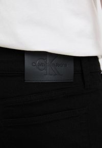A fekete farmer nadrágon egy textúrázott fekete bőr folt található, amelyen a "Calvin Klein Jeans" logó és a "CK" kezdőbetűk vannak bemélyítve, és a deréknál van rögzítve.