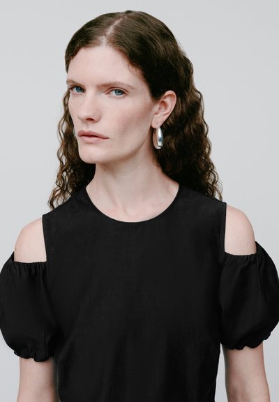 Frau mit langen, lockigen braunen Haaren, die silberne Creolen und ein schwarzes Top mit freier Schulter trägt, schaut leicht zur Seite.