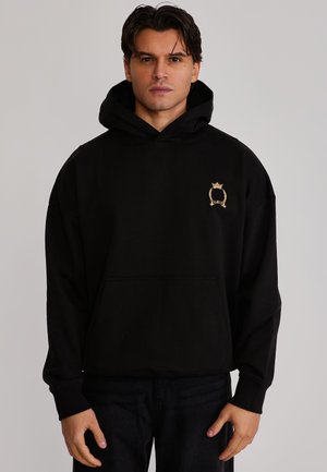 Homme portant un sweat à capuche noir avec un petit emblème doré brodé sur le côté gauche de la poitrine, debout devant un fond uni.