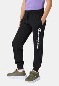 Pantaloni da jogging neri con vita elastica e tasche laterali, con il logo bianco "Champion" stampato lungo la gamba sinistra.