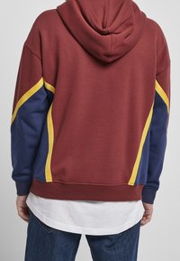 Sweat à capuche couleur bordeaux avec des accents bleu marine et jaune. Matière en coton, coupe décontractée, poignets et ourlet côtelés. Superposé avec une chemise blanche en dessous.