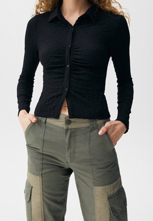 Femme portant une chemise noire cintrée à boutons et froncée, et un pantalon cargo bicolore vert, les mains dans les poches, cadrée à mi-torse.