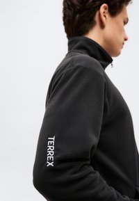 Jeune personne portant une veste polaire noire avec un col haut, logo "TERREX" brodé sur la manche gauche, faisant face à droite.