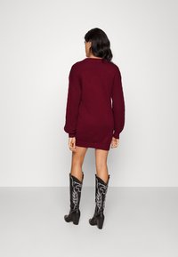 Donna con lunghi capelli scuri, vestita con un abito maglione bordeaux e stivali neri in stile western con tacco, in piedi di fronte a un muro bianco.