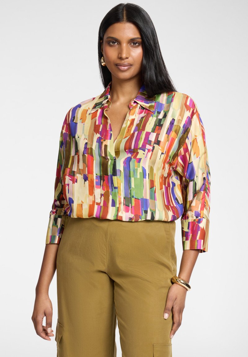 Mujer con cabello largo y oscuro, vistiendo una blusa de estampado abstracto multicolor y pantalones anchos color mostaza, posando con una mano relajada.