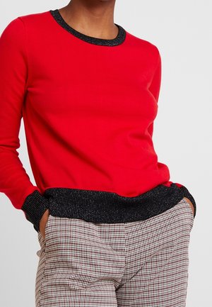 Personne portant un haut rouge à manches longues avec bordure noire et un pantalon à carreaux taille haute, les mains dans les poches, sur un fond uni.