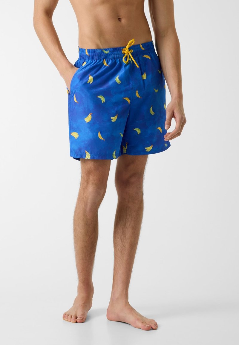 Homme portant un short de bain bleu avec des imprimés bananes jaunes et un cordon jaune, debout pieds nus avec une main dans la poche.