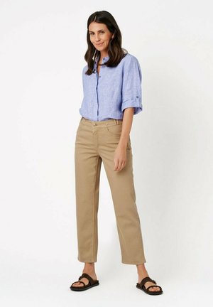 Frau mit langen dunklen Haaren, die ein hellblaues Hemd mit Knöpfen, beige gerade geschnittene Hosen und schwarze Sandalen trägt, steht vor einem weißen Hintergrund.