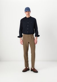 Camicia blu con bottoni, pantaloni beige su misura e mocassini marroni. Un berretto blu aggiunge un tocco di dettaglio. Il modello è in piedi contro uno sfondo bianco.