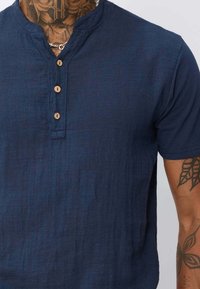 Chemise henley bleu marine à manches courtes avec un tissu texturé, comportant trois boutons en bois et un col sans col.