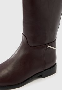 Σκούρο καφέ δερμάτινο μπότες έως τη μέση της γάμπας με χαμηλό μαύρο τακούνι και ασημένια λεπτομέρεια σε μορφή δαχτυλιδιού στο εξωτερικό μέρος κοντά στον αστράγαλο.