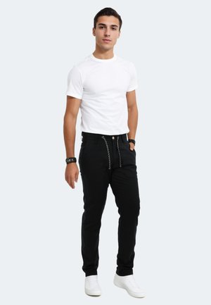 Schwarze Hose mit strukturiertem Muster und dekorativen Kettendetails, kombiniert mit einem schlichten weißen T-Shirt und weißen Sneakers.