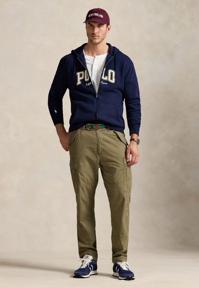 Marinblå zip-up hoodie med "POLO" logo, beige cargobyxor, vinröd keps och marinblå sneakers. Har en avslappnad passform och ytterligare fickdetaljer.