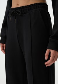 Zwarte sweatpants van zacht materiaal, met een elastische tailleband met een koord, zijzakken en een losse pasvorm aan de benen.