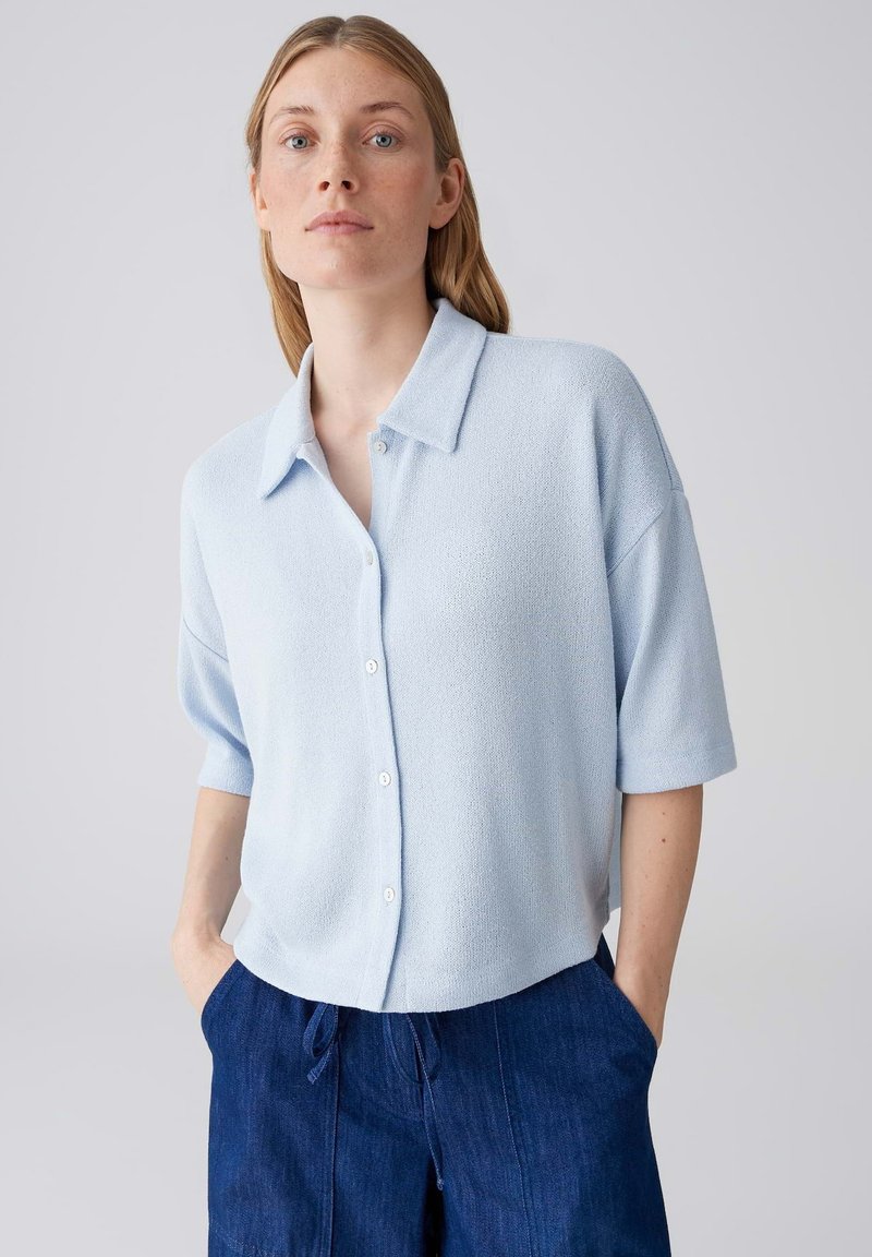 Donna con capelli biondi che indossa una camicia celeste a maniche corte con bottoni e pantaloni blu scuro, con le mani nelle tasche, sfondo neutro.