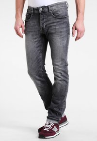 Hombre vestido con jeans ajustados negros desgastados y zapatillas maroon de New Balance, de pie con las piernas cruzadas.