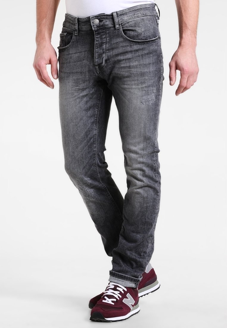 Hombre vestido con jeans ajustados negros desgastados y zapatillas maroon de New Balance, de pie con las piernas cruzadas.