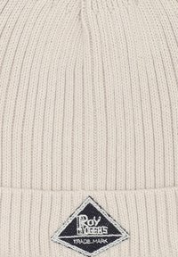 Berretto lavorato a maglia di colore crema morbido con texture a costine. Presenta una toppa a forma di diamante nera con testo e logo bianchi, posizionata sul risvolto.