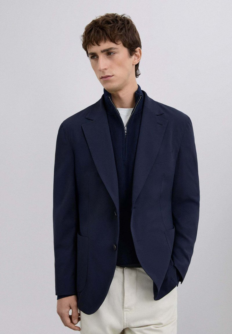 Blazer navy de corte entallado, con solapas de pico, dos botones y bolsillos laterales, usado sobre una blusa de punto navy y pantalones de color claro.