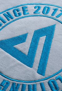 Logo géométrique brodé en bleu avec le texte "DEPUIS 2017" sur un tissu gris clair, montrant une texture de couture détaillée.