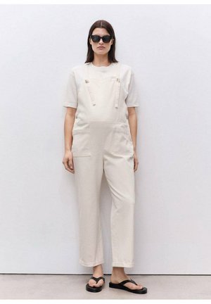 Cream-colored overall with loose fit, featuring adjustable straps, two front pockets, and paired with a short-sleeve top and flip-flops.