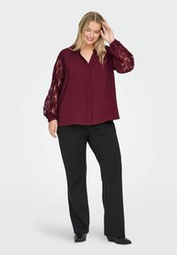 Blusa bordeaux con bottoni e maniche a palloncino in pizzo trasparente; abbinata a pantaloni neri a gamba dritta e scarpe nere con tacco.