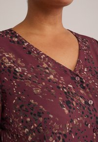 Blusa bordeaux con scollatura a V, caratterizzata da un motivo a macchie in beige, nero e oro, arricchita da piccoli bottoni lungo il davanti.