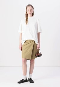 Biela oversized tričko, olivová sukňa s plisovaním, model nosí čierne mokasíny a biele ponožky, drží malú hnedú kabelku.