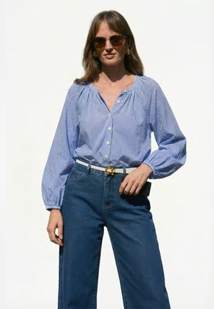 Vrouw met schildpadkleurige zonnebril, gouden oorbellen, blauw geruit blouse met knopen, hooggetailleerde blauwe jeans en witte riem tegen een witte achtergrond.