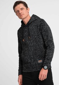 Schwarze und graue gesprenkelte Kapuzenjacke mit Kordelzugkapuze, lederähnlichem Akzent und gerippten Bündchen, getragen mit schwarzen Hosen und einer Armbanduhr.