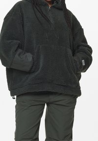 Felpa con cappuccio fuzzy verde scuro, con mezza zip, vestibilità oversize, tasca frontale e polsini a costine. Abbinata a pantaloni verdi coordinati.
