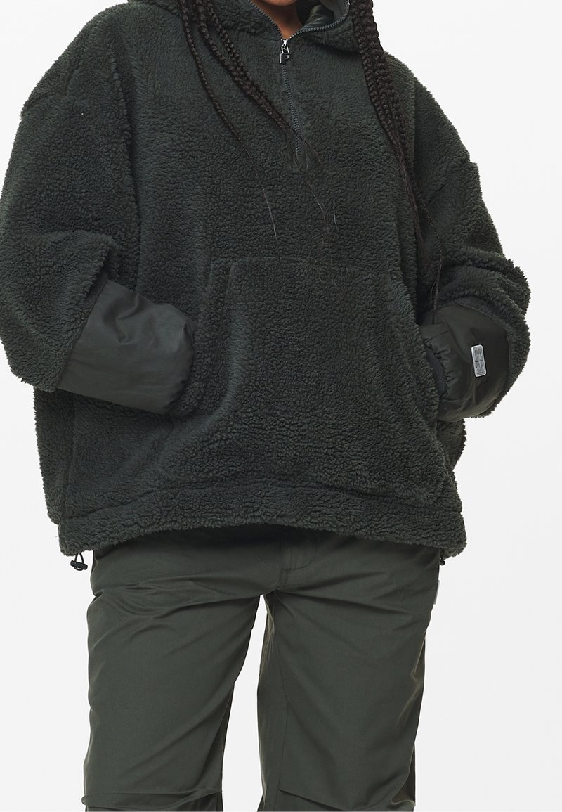 Felpa con cappuccio fuzzy verde scuro, con mezza zip, vestibilità oversize, tasca frontale e polsini a costine. Abbinata a pantaloni verdi coordinati.