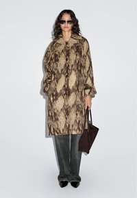 Braun- und beiges, gemustertes, übergroßes Mäntel mit Kragen und Knöpfen, kombiniert mit dunkelblauen Jeans und spitzen schwarzen Schuhen, während eine dunkle Tasche gehalten wird.