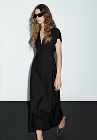 Svart knälång klänning med V-ringning, korta ärmar och en figurnära silhuett. Tillverkad av ett mjukt tyg, i kombination med grå, spetsiga skor.